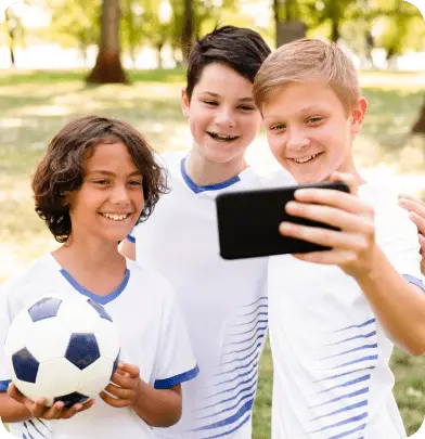 Three young soccer players smiling while taking a selfie, one holding a soccer ball.